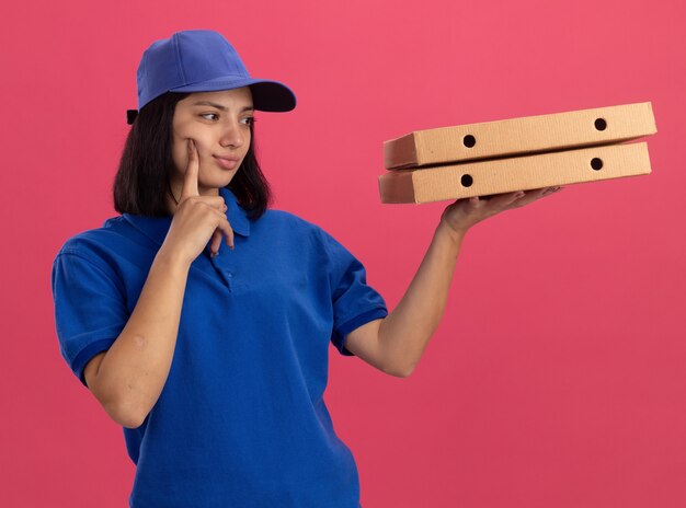 Repartidor joven en uniforme azul y gorra sosteniendo cajas de pizza mirándolos con expresión pensativa pensando de pie sobre la pared rosa