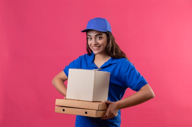 Repartidor joven en uniforme azul y gorra sosteniendo cajas de pizza y paquete de caja mirando a la cámara sonriendo alegremente feliz y positivo de pie sobre fondo rosa