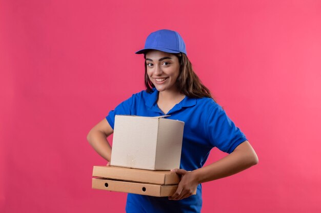 Repartidor joven en uniforme azul y gorra sosteniendo cajas de pizza y paquete de caja mirando a la cámara sonriendo alegremente feliz y positivo de pie sobre fondo rosa