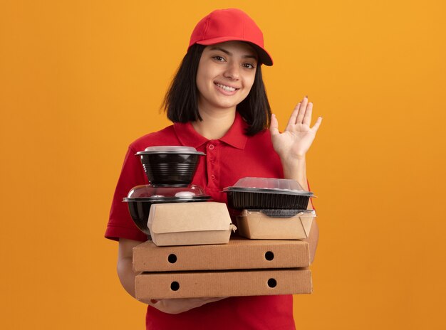Repartidor joven en uniforme azul y gorra sosteniendo cajas de pizza y paquete de comida sonriendo feliz y positivo saludando con la mano de pie sobre la pared naranja