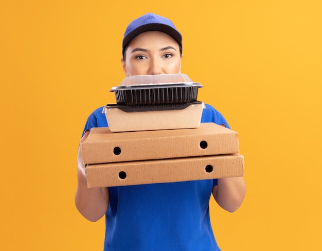 Repartidor joven en uniforme azul y gorra sosteniendo cajas de pizza y paquetes de alimentos mirando al frente con cara seria de pie sobre la pared naranja