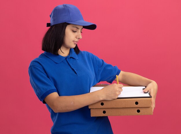 Repartidor joven en uniforme azul y gorra sosteniendo cajas de pizza y portapapeles escribiendo algo con cara seria de pie sobre la pared rosa