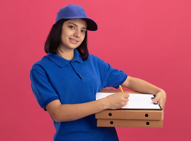 Repartidor joven en uniforme azul y gorra sosteniendo cajas de pizza y portapapeles con lápiz con sonrisa en la cara de pie sobre la pared rosa