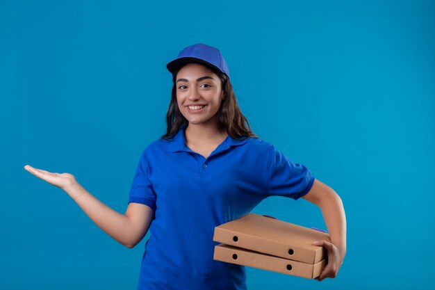 Repartidor joven en uniforme azul y gorra sosteniendo cajas de pizza presentando con el brazo de la mano sonriendo alegremente de pie sobre fondo azul.