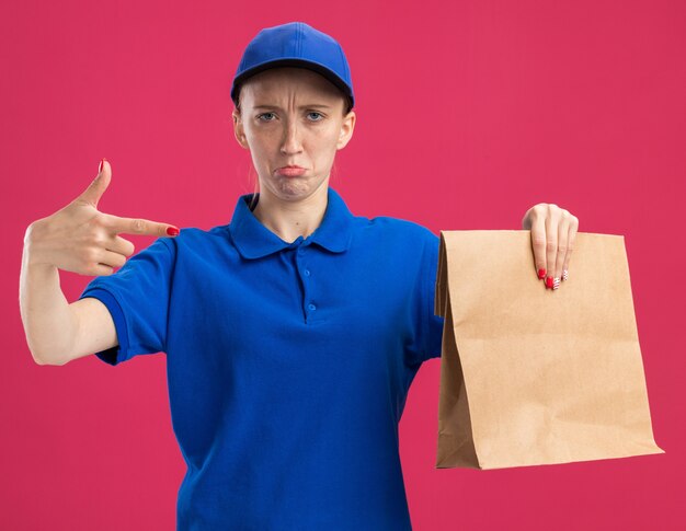 Repartidor joven en uniforme azul y gorra sosteniendo el paquete de papel apuntando con el dedo índice hacia él con expresión triste frunciendo los labios de pie sobre la pared rosa