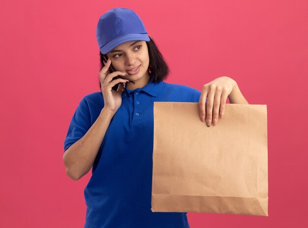 Repartidor joven en uniforme azul y gorra sosteniendo el paquete de papel hablando por teléfono móvil con una sonrisa en la cara de pie sobre la pared rosa