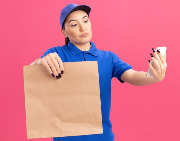 Repartidor joven en uniforme azul y gorra sosteniendo el paquete de papel mirando su teléfono inteligente con cara seria de pie sobre la pared rosa