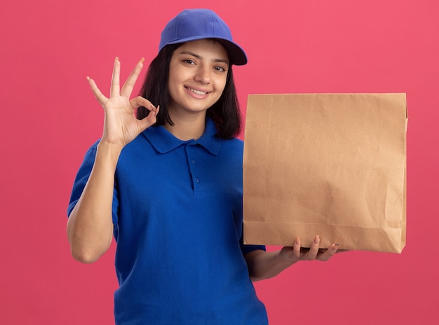 Repartidor joven en uniforme azul y gorra sosteniendo el paquete de papel mostrando el signo de ok sonriendo alegremente de pie sobre la pared rosa