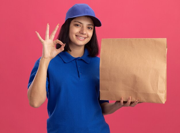 Repartidor joven en uniforme azul y gorra sosteniendo el paquete de papel mostrando el signo de ok sonriendo alegremente de pie sobre la pared rosa