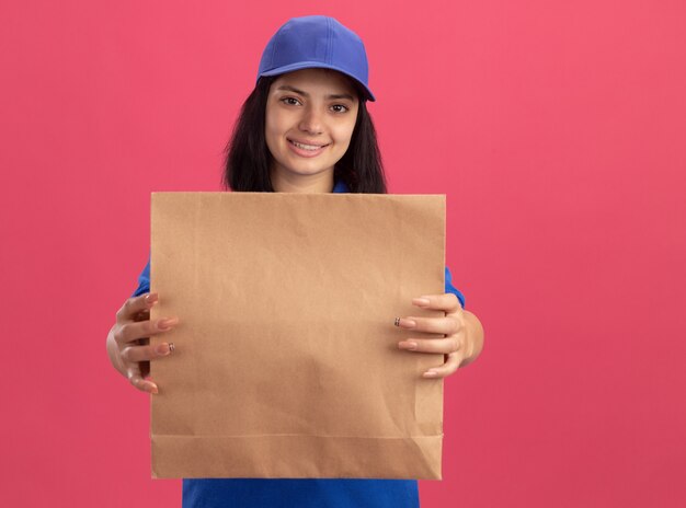 Repartidor joven en uniforme azul y gorra sosteniendo el paquete de papel sonriendo confiado parado sobre la pared rosa