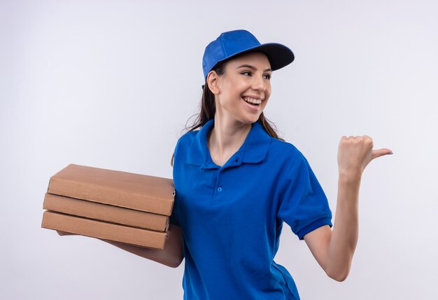 Repartidor joven en uniforme azul y gorra sosteniendo la pila de cajas de pizza apuntando hacia atrás con el pulgar sonriendo alegremente