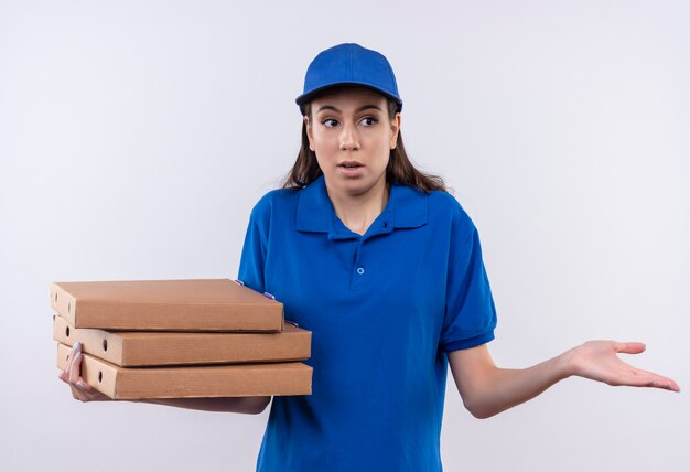 Repartidor joven en uniforme azul y gorra sosteniendo una pila de cajas de pizza mirando incierto y confundido encogiéndose de hombros