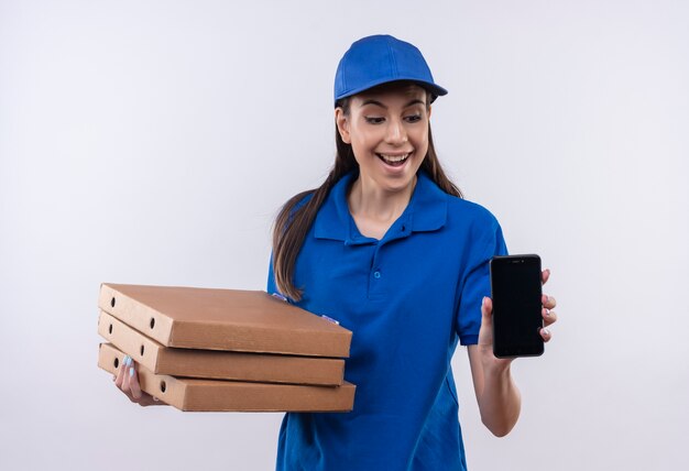 Repartidor joven en uniforme azul y gorra sosteniendo la pila de cajas de pizza mostrando smartphone sonriendo alegremente