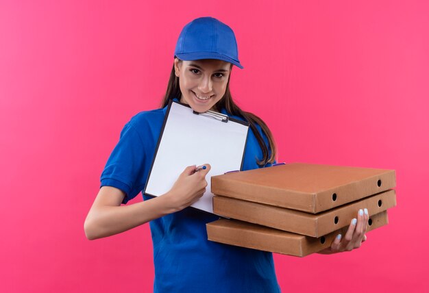 Repartidor joven en uniforme azul y gorra sosteniendo una pila de cajas de pizza y portapapeles con páginas en blanco pidiendo firma