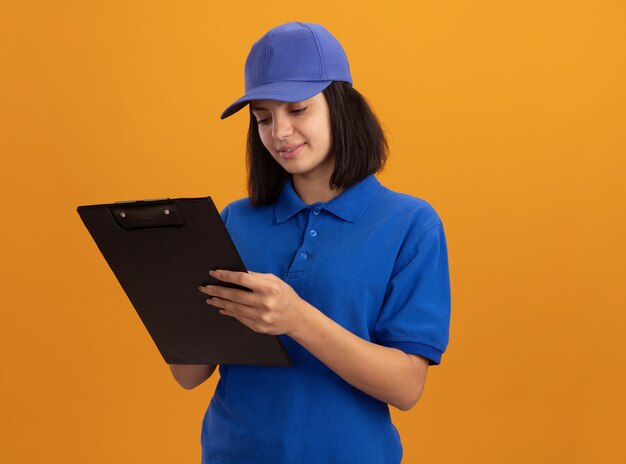 Repartidor joven en uniforme azul y gorra sosteniendo portapapeles mirándolo con una sonrisa en la cara de pie sobre la pared naranja