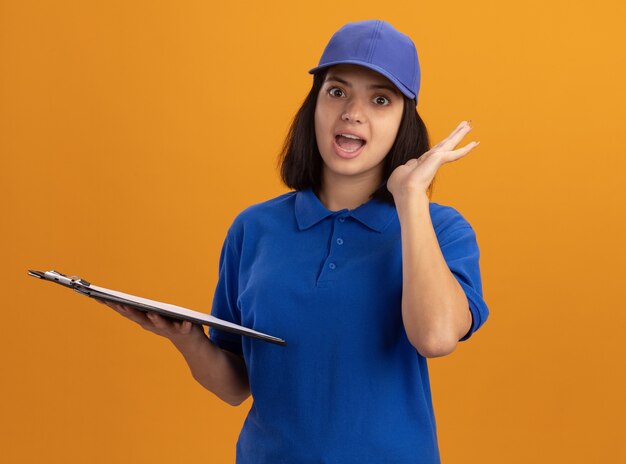 Repartidor joven en uniforme azul y gorra sosteniendo el portapapeles sonriendo sorprendido de pie sobre la pared naranja