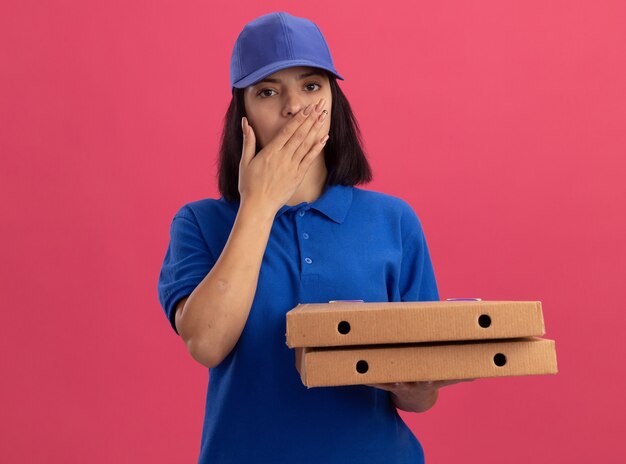 Repartidor joven en uniforme azul sosteniendo cajas de pizza sorprendido cubriendo la boca con la mano de pie sobre la pared rosa