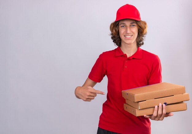 Repartidor joven en uniforme rojo apuntando a cajas de pizza en su mano mirando a la cámara sonriendo alegremente sobre fondo blanco aislado