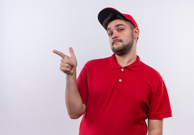 Repartidor joven en uniforme rojo y gorra apuntando con el dedo hacia el lado mirando confiado