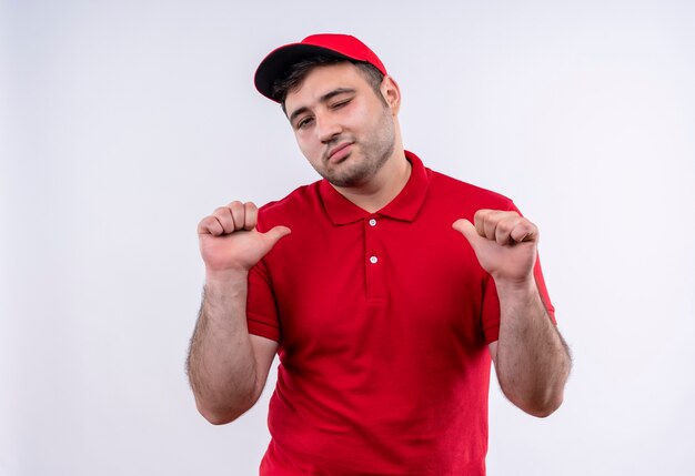 Repartidor joven con uniforme rojo y gorra apuntando con los pulgares a sí mismo, satisfecho de sí mismo y orgulloso de pie sobre la pared blanca