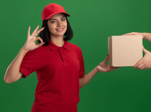 Repartidor joven en uniforme rojo y gorra dando caja de cartón a un cliente sonriendo amable mostrando signo ok de pie sobre la pared verde