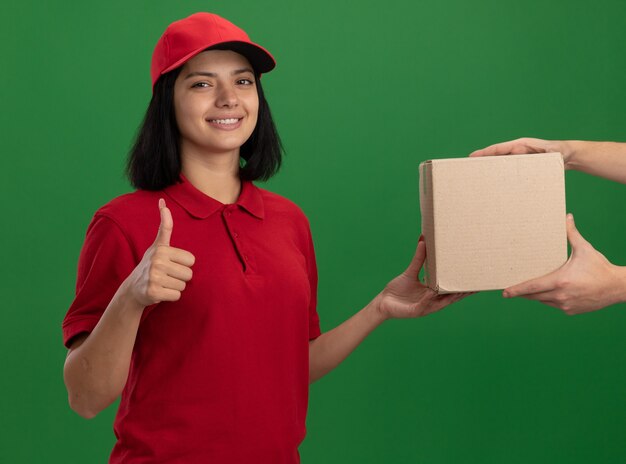 Repartidor joven en uniforme rojo y gorra dando caja de cartón a un cliente sonriendo amigable mostrando los pulgares para arriba de pie sobre la pared verde