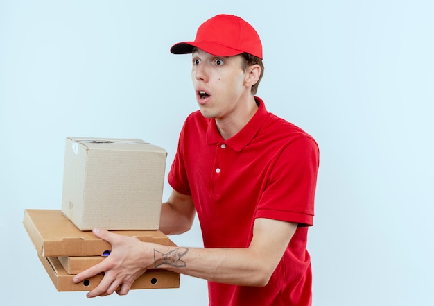 Repartidor joven en uniforme rojo y gorra dando paquetes de caja a un cliente que parece sorprendido de pie sobre una pared blanca