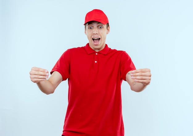 Repartidor joven en uniforme rojo y gorra emocionado y feliz gesticulando con las manos, concepto de lenguaje corporal de pie sobre una pared blanca