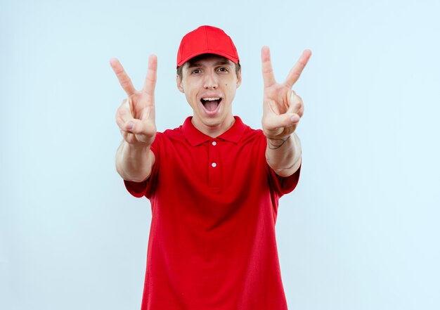 Repartidor joven en uniforme rojo y gorra feliz y emocionado mostrando el signo de la victoria con ambas manos de pie sobre la pared blanca