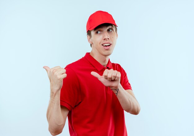 Repartidor joven en uniforme rojo y gorra feliz y positiva apuntando hacia atrás con los pulgares de pie sobre la pared blanca