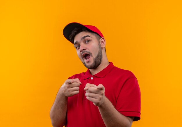 Repartidor joven en uniforme rojo y gorra feliz y positiva apuntando con los dedos a la cámara