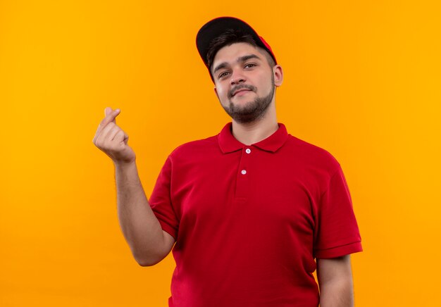 Repartidor joven en uniforme rojo y gorra frotando los dedos haciendo gesto de dinero pidiendo dinero