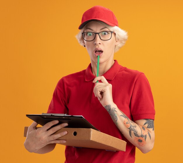 Repartidor joven en uniforme rojo y gorra con gafas con caja de pizza y portapapeles y lápiz de pie preocupado y confundido sobre la pared naranja