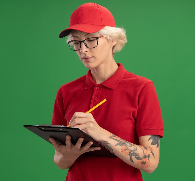 Repartidor joven en uniforme rojo y gorra con gafas con portapapeles con páginas en blanco y lápiz escribiendo algo con cara seria de pie sobre la pared verde