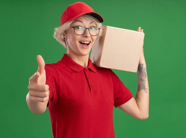 Repartidor joven en uniforme rojo y gorra con gafas sosteniendo una caja de cartón mirando feliz y emocionado mostrando los pulgares para arriba de pie sobre la pared verde