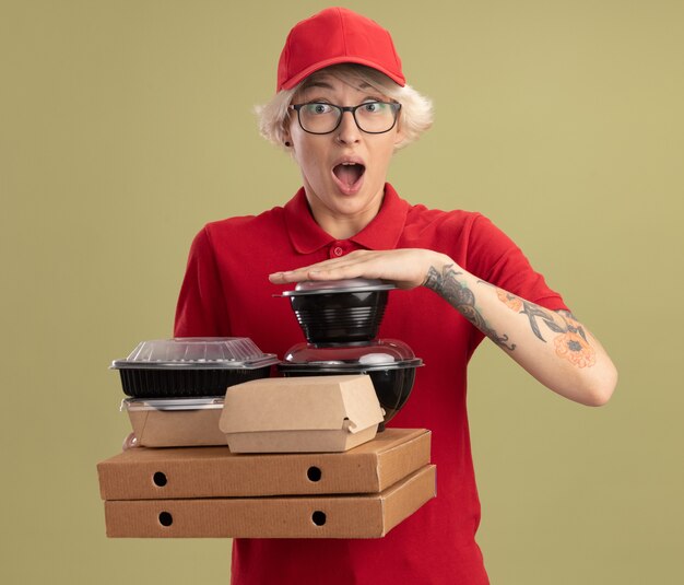 Repartidor joven en uniforme rojo y gorra con gafas sosteniendo cajas de pizza y paquetes de alimentos feliz y sorprendido de pie sobre la pared verde
