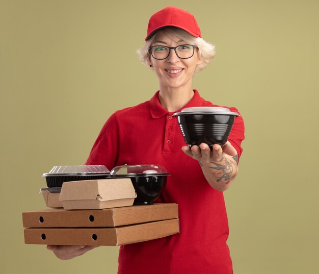 Repartidor joven en uniforme rojo y gorra con gafas sosteniendo cajas de pizza y paquetes de alimentos sonriendo alegremente ofreciendo caja de pie sobre la pared verde