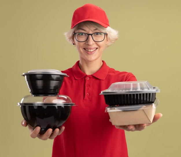 Repartidor joven en uniforme rojo y gorra con gafas sosteniendo paquetes de alimentos con una sonrisa en la cara de pie sobre la pared verde
