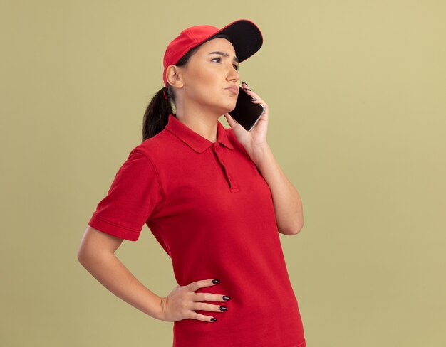 Repartidor joven en uniforme rojo y gorra hablando por teléfono móvil mirando confundido de pie sobre la pared verde