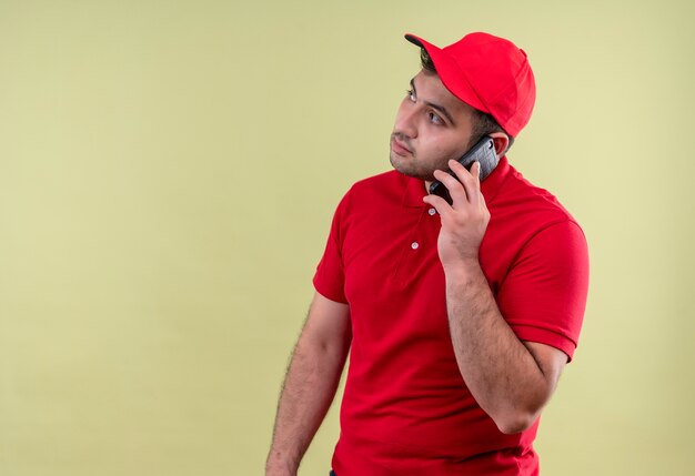 Repartidor joven en uniforme rojo y gorra hablando por teléfono móvil mirando a un lado con cara seria de pie sobre la pared verde