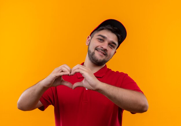 Foto gratuita repartidor joven en uniforme rojo y gorra haciendo gesto de corazón con los dedos sobre el pecho sonriendo