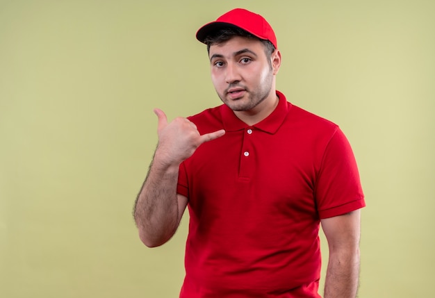 Foto gratuita repartidor joven en uniforme rojo y gorra haciendo gesto de llamarme sonriendo confiado de pie sobre la pared verde