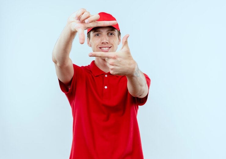 Foto gratuito repartidor joven en uniforme rojo y gorra haciendo marco con los dedos sonriendo mirando hacia el frente a través de este marco parado sobre la pared blanca