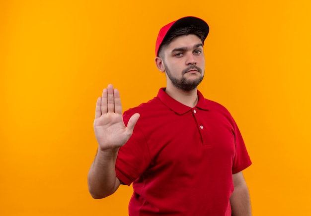 Repartidor joven en uniforme rojo y gorra haciendo señal de pare con la mano mirando con cara seria