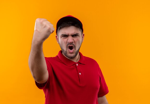 Repartidor joven en uniforme rojo y gorra levantando el puño mostrando a la cámara con cara enojada