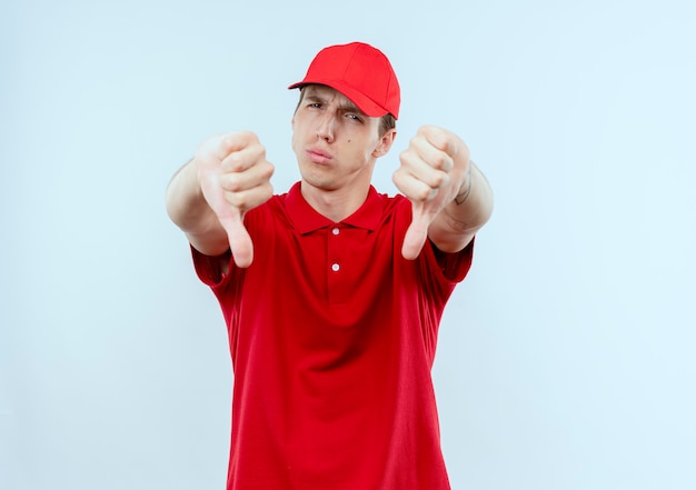 Foto gratuita repartidor joven con uniforme rojo y gorra mirando al frente disgustado mostrando los pulgares hacia abajo de pie sobre la pared blanca