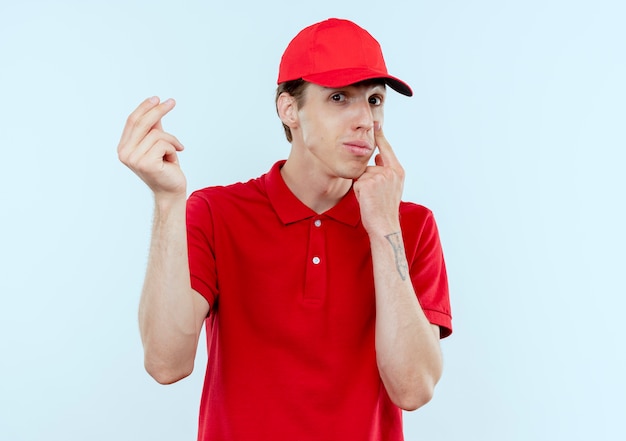 Foto gratuita repartidor joven con uniforme rojo y gorra mirando al frente frotándose los dedos pidiendo dinero apuntando con el dedo a su ojo esperando el pago de pie sobre una pared blanca