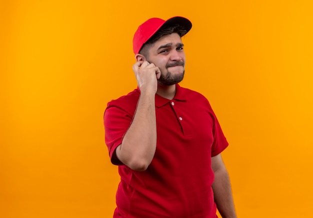 Repartidor joven en uniforme rojo y gorra mirando a cámara con expresión escéptica en la cara tocando la oreja