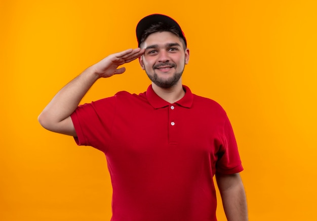 Foto gratuita repartidor joven en uniforme rojo y gorra mirando confiado saludando