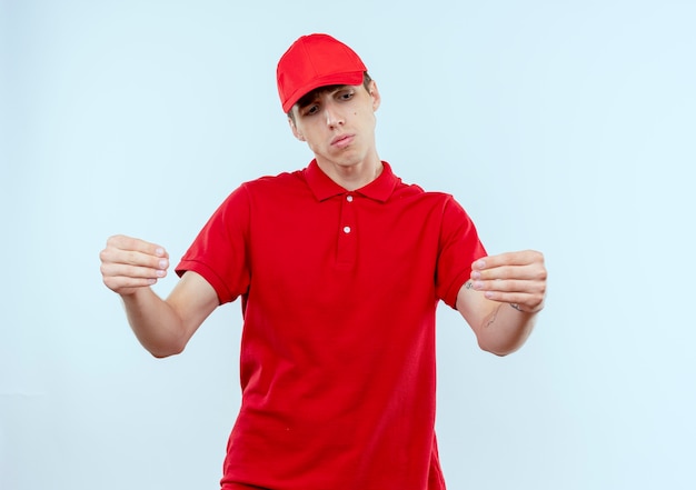 Repartidor joven en uniforme rojo y gorra mirando con expresión triste en la cara haciendo gestos con las manos, concepto de lenguaje corporal de pie sobre una pared blanca